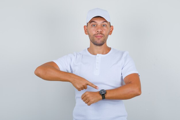 Young man in white t-shirt, cap showing watch with index finger and looking excited, front view.