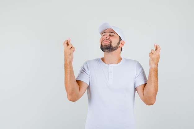 Young man in white t-shirt,cap standing with crossed fingers and looking confident , front view.