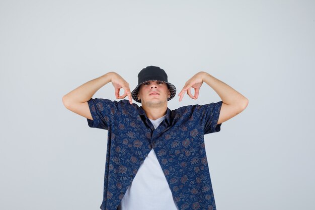 Young man in white t-shirt, floral shirt, cap showing ok signs and looking confident , front view.