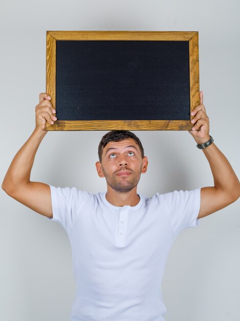 Young man in white t-shirt holding blackboard above his head, front view.