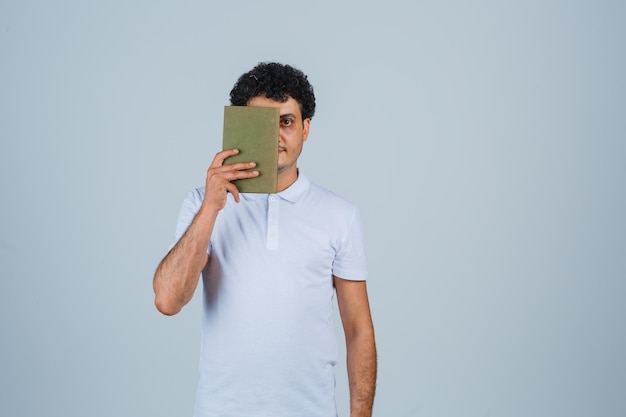 Young man in white t-shirt holding book and looking cheerful , front view.