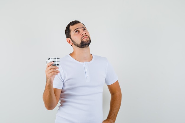 Young man in white t-shirt holding cup while looking away and looking thoughtful