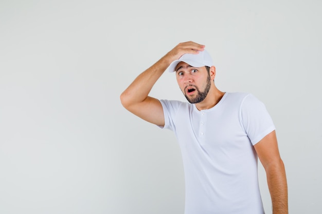 Young man in white t-shirt holding hand on his head and looking scared , front view.