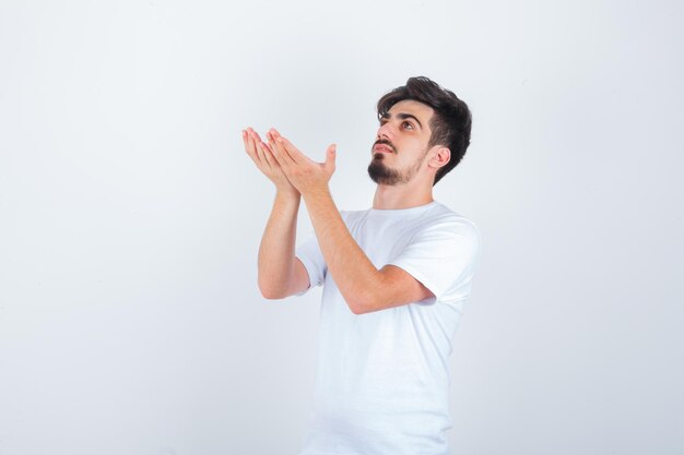 Young man in white t-shirt holding hands in praying gesture and looking hopeful