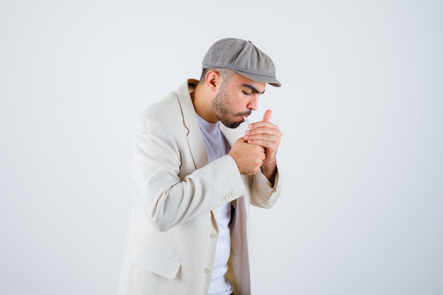 Молодой человек в белой футболке, куртке и серой кепке курит сигареты и выглядит сосредоточенным