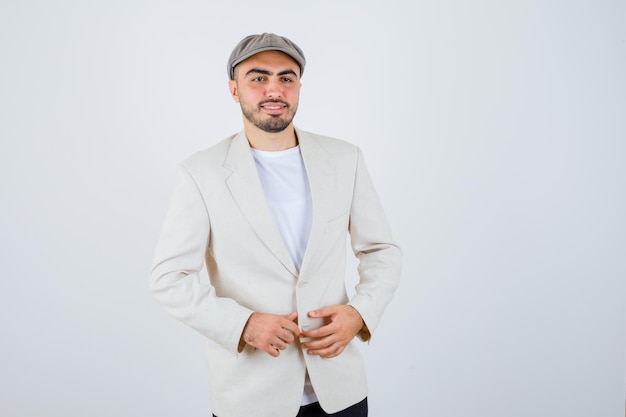 Free photo young man in white t-shirt, jacket and grey cap holding hands on jacket and looking happy