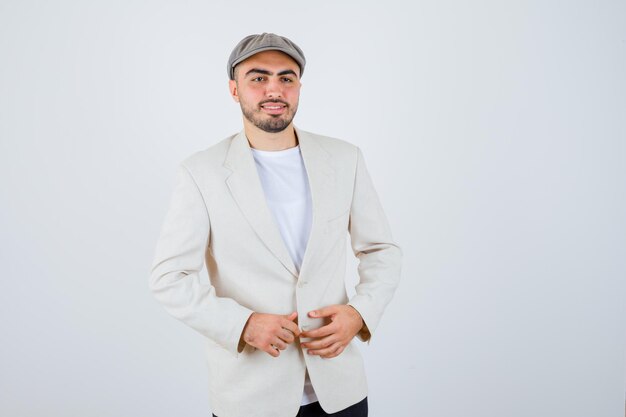 Young man in white t-shirt, jacket and grey cap holding hands on jacket and looking happy