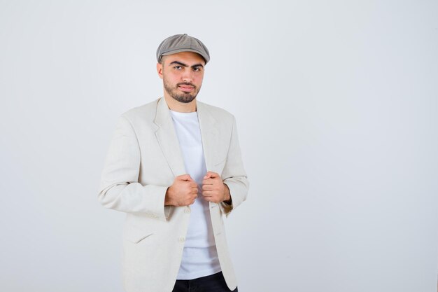 Young man in white t-shirt, jacket and grey cap holding hands in jacket and looking serious