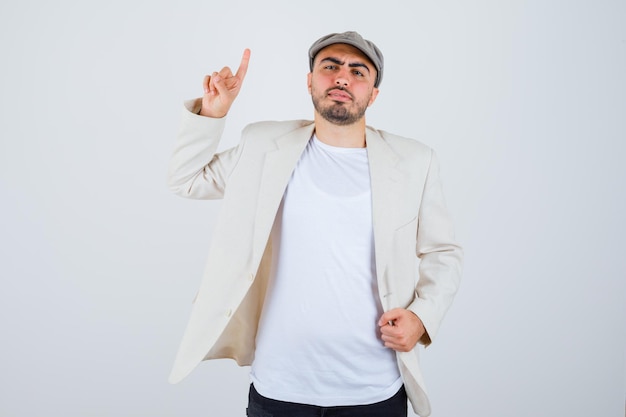 Young man in white t-shirt, jacket and grey cap raising index finger in eureka gesture and holding hand on jacket and looking serious