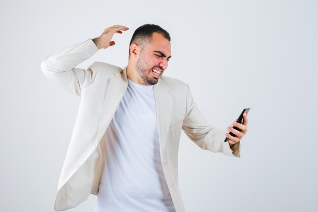 Young man in white t-shirt, jacket holding mobile phone and looking at it and looking furious , front view.