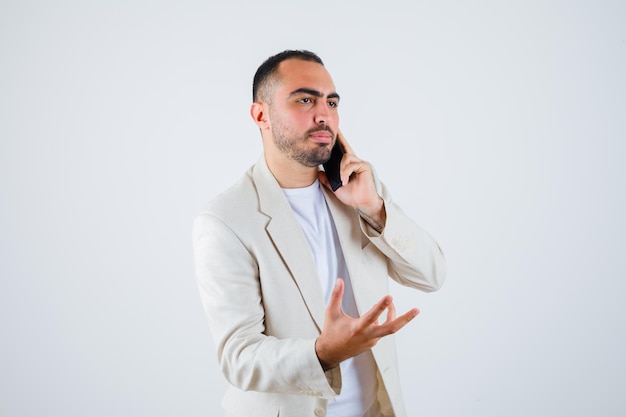 Young man in white t-shirt, jacket talking to phone and stretching hand as holding something and looking serious , front view.