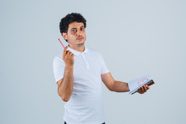 Young man in white t-shirt and jeans biting pen, holding notebook and thinking about something and looking pensive , front view.