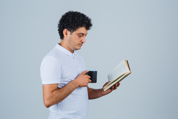 흰색 티셔츠와 청바지를 입은 젊은 남자는 책을 읽고 집중된 정면을 바라보면서 차 한 잔을 마시고 있습니다.