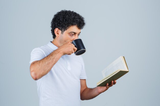 Young man in white t-shirt and jeans drinking cup of tea while reading book and looking happy , front view.