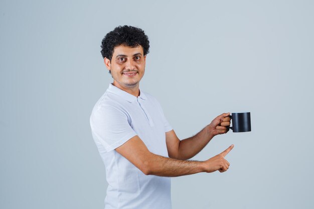 Молодой человек в белой футболке и джинсах держит чашку чая, указывая на него и выглядит счастливым, вид спереди.