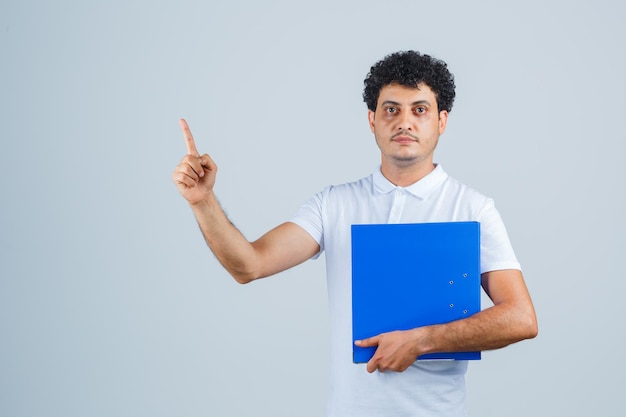 Молодой человек в белой футболке и джинсах держит папку с файлами, поднимает указательный палец в жесте эврики и выглядит разумно, вид спереди.