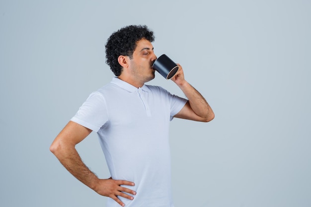 Young man in white t-shirt and jeans holding hand on waist while drinking cup of tea and looking thirsty , front view.