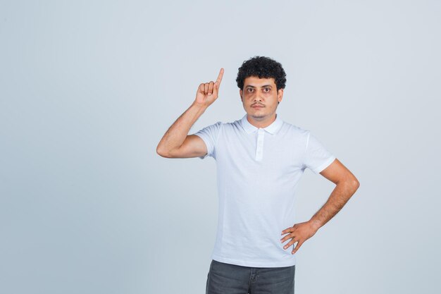 Молодой человек в белой футболке и джинсах держит руку на талии, поднимая указательный палец в жесте эврики и выглядит разумно, вид спереди.