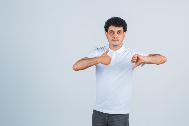 Young man in white t-shirt and jeans showing thumbs up and down with both hands and looking serious , front view.