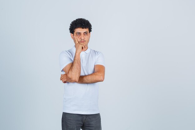 Young man in white t-shirt and jeans standing in thinking pose, leaning cheek on hand and looking pensive , front view.