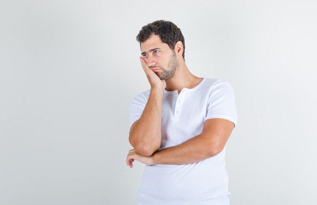 Young man in white t-shirt leaning his cheek on raised palm and looking thoughtful