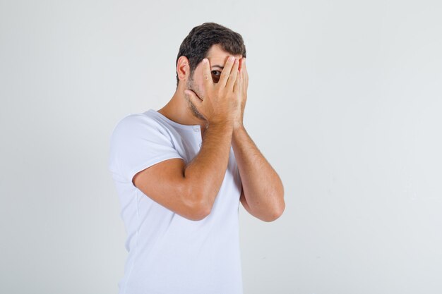 Young man in white t-shirt looking through fingers with one eye