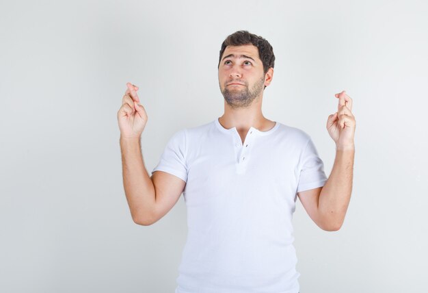 Young man in white t-shirt looking up with crossed fingers and looking hopeful