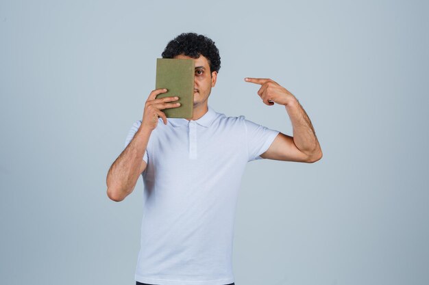 Young man in white t-shirt pointing at book and looking confident , front view.