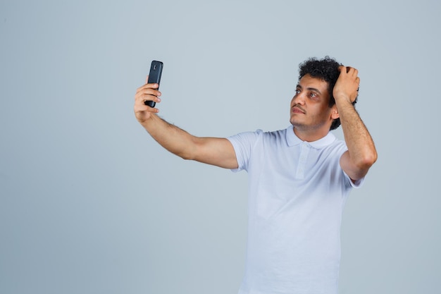 Young man in white t-shirt posing while taking selfie and looking cute , front view.