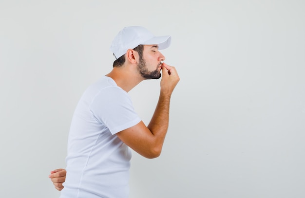 Young man in white t-shirt showing delicious gesture and looking focused , front view.