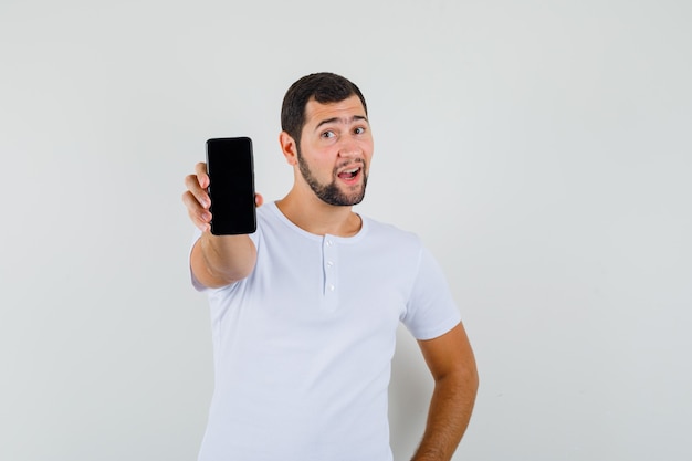 Молодой человек в белой футболке показывает мобильный телефон и выглядит счастливым, вид спереди.