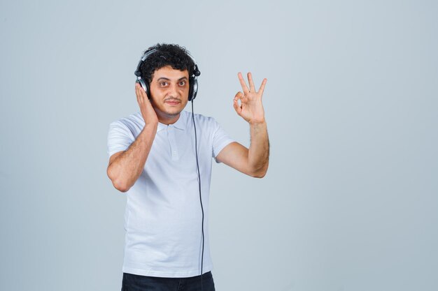 Young man in white t-shirt showing ok gesture while listening to music and looking confident , front view.