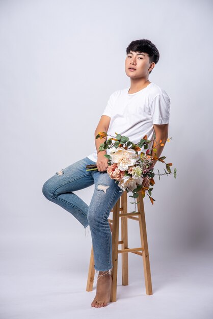 A young man in a white t-shirt sits on a high chair and holds flowers.