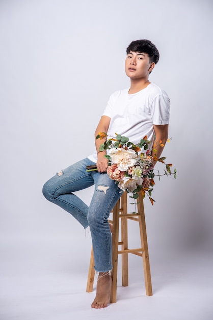 Молодой человек в белой футболке сидит на стульчике и держит цветы.