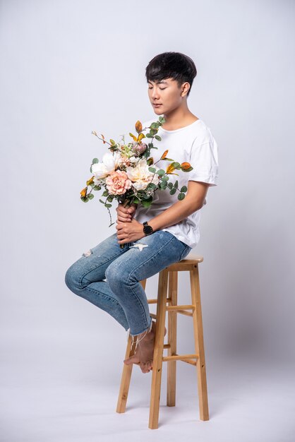 A young man in a white t-shirt sits on a high chair and holds flowers.