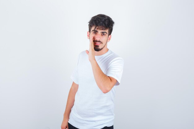 Young man in white t-shirt telling secret behind hand and looking confident