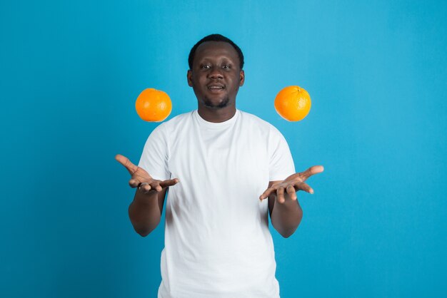 Young man in white t-shirt throwing up two sweet orange fruits against blue wall 