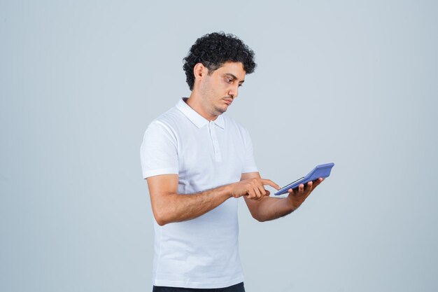 Young man in white t-shirt using calculator and looking pensive , front view.