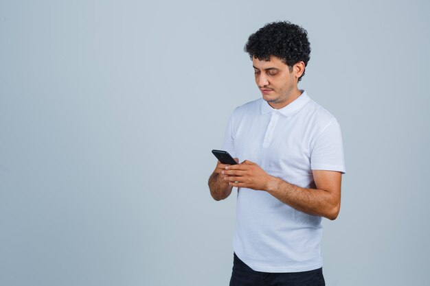Молодой человек в белой футболке с помощью мобильного телефона и выглядит занятым, вид спереди.