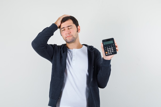 Young man in white t-shirt and zip-front black hoodie holding calculator and putting hand on head and looking optimistic , front view.