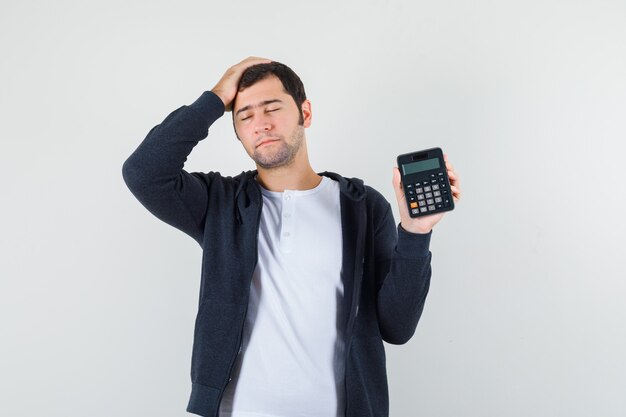 白いTシャツとジッパー付きの若い男-電卓を保持し、頭に手を置いて、楽観的な正面図を探しているフロントの黒いパーカー。