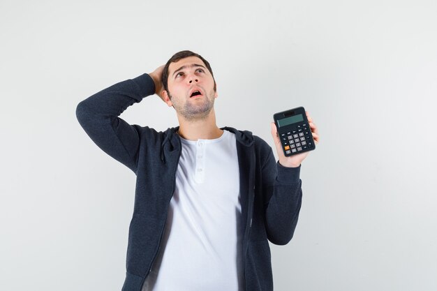 Young man in white t-shirt and zip-front black hoodie holding calculator and putting hand on head and looking pensive , front view.