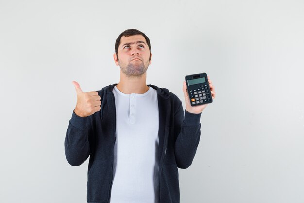 Молодой человек в белой футболке и черной толстовке с капюшоном на молнии, держит калькулятор и показывает большой палец вверх и задумчиво смотрит, вид спереди.