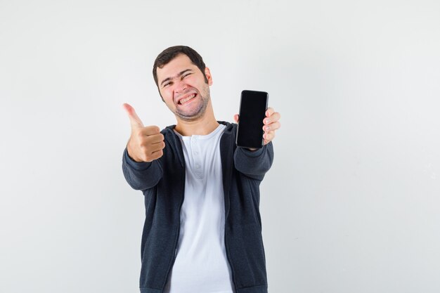 Молодой человек в белой футболке и черной толстовке с капюшоном на молнии, держа смартфон, показывает большой палец вверх и выглядит счастливым, вид спереди.