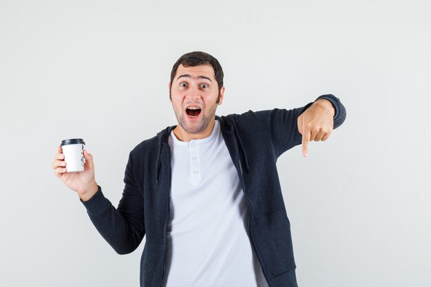 Young man in white t-shirt and zip-front black hoodie holding takeaway coffee cup and pointing down with index finger and looking surprised , front view.