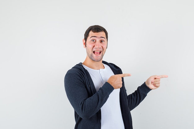 Young man in white t-shirt and zip-front black hoodie pointing to right with index fingers and looking optimistic , front view.