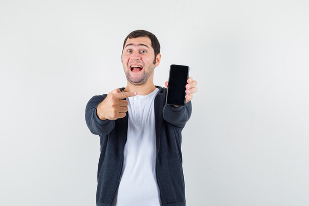 Young man in white t-shirt and zip-front black hoodie pointing smartphone with index finger and looking happy , front view.