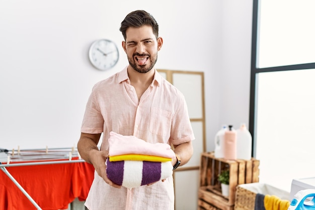 Young man with beard holding folded laundry after ironing sticking tongue out happy with funny expression