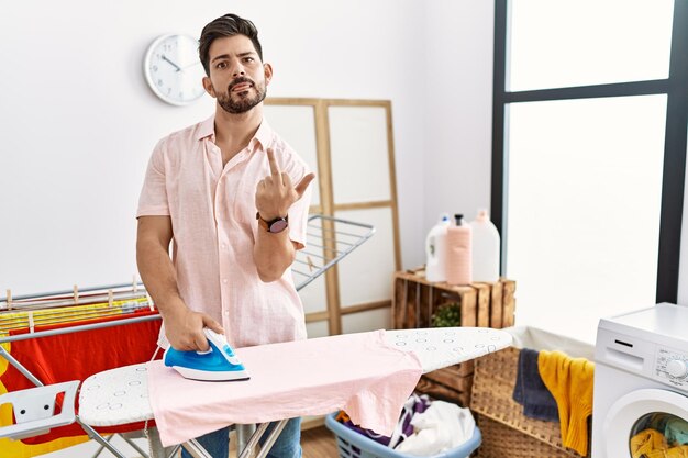 Free photo young man with beard ironing clothes at home showing middle finger impolite and rude fuck off expression