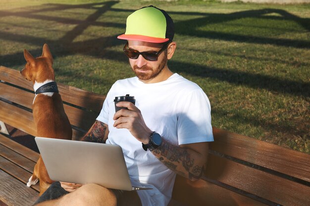 Young man with beard and tattoos wearing a plain white t-shirt drinking coffee and looking at his laptop while his brown and white dog sits next to him on a park bench.